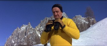 Movie still from “The Pink Panther” (1963), directed by Blake Edwards – A man in a yellow sweater holding a pair of binoculars in front of a mountain range; Medium shot, Low angle
