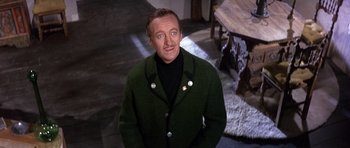 Movie still from “The Pink Panther” (1963), directed by Blake Edwards – A man in a green coat standing next to a wooden table; Medium shot, High angle