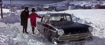Movie still from “The Pink Panther” (1963), directed by Blake Edwards – A woman in a red coat is standing next to an old car in the snow; Wide shot, High angle