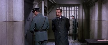 Movie still from “The Pink Panther” (1963), directed by Blake Edwards – A man in a trench coat standing next to another man in a uniform; Medium shot, Low angle