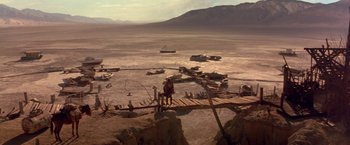 Movie still from “The Postman” (1997), directed by Kevin Costner – A man standing on a wooden bridge in the middle of the desert; Extreme Wide shot, High angle