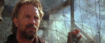Movie still from “The Postman” (1997), directed by Kevin Costner – A man with a beard is looking through a wire fence; Close Up shot, Over the shoulder angle