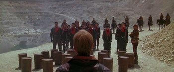 Movie still from “The Postman” (1997), directed by Kevin Costner – A group of men in black and red outfits standing in front of a man; Extreme Wide shot, High angle