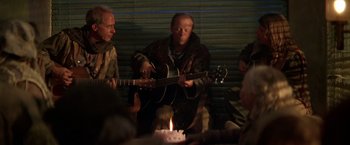 Movie still from “The Postman” (1997), directed by Kevin Costner – A group of people sitting around a table with a guitar; Medium shot, Over the shoulder angle