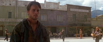 Movie still from “The Postman” (1997), directed by Kevin Costner – A man standing in front of a building on a city street; Medium shot, Over the shoulder angle