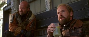 Movie still from “The Postman” (1997), directed by Kevin Costner – A man sitting next to another man holding a jar; Close Up shot, Low angle