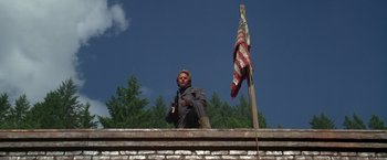 Movie still from “The Postman” (1997), directed by Kevin Costner – A man sitting on top of a brick building next to an american flag; Wide shot, Low angle