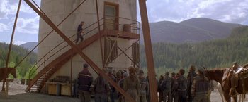 Movie still from “The Postman” (1997), directed by Kevin Costner – A group of people standing around a building; Extreme Wide shot, High angle