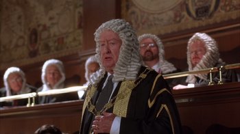 Movie still from “The Princess Diaries 2: Royal Engagement” (2004), directed by Garry Marshall – A man in a judge's wig and wig stand in front of judges; Medium shot, Over the shoulder angle