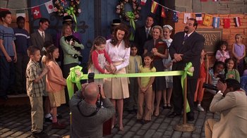Movie still from “The Princess Diaries 2: Royal Engagement” (2004), directed by Garry Marshall – A woman cutting a green ribbon with a man taking a picture of it; Wide shot, High angle