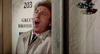 Movie still from “The Producers” (1967), directed by Mel Brooks – A man in a trench coat and a tie is standing next to a wall; Close Up shot, Low angle