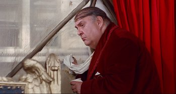 Movie still from “The Producers” (1967), directed by Mel Brooks – A man in a red jacket is holding a cup; Medium shot, High angle