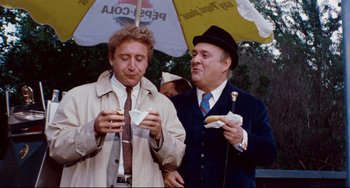Movie still from “The Producers” (1967), directed by Mel Brooks – A couple of men standing next to each other under an umbrella; Medium shot, Over the shoulder angle