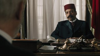 Movie still from “The Promise” (2016), directed by Terry George – A man sitting at a table wearing a red fez; Medium shot, Over the shoulder angle