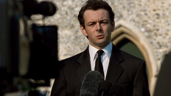 Movie still from “The Queen” (2006), directed by Stephen Frears – A man wearing a suit and tie standing next to a microphone; Close Up shot, Over the shoulder angle