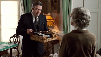 Movie still from “The Queen” (2006), directed by Stephen Frears – An older man in a suit and tie holding a basket of papers; Medium shot, Over the shoulder angle
