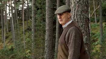 Movie still from “The Queen” (2006), directed by Stephen Frears – An older man wearing a hat and vest in a wooded area; Medium shot, High angle