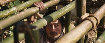 Movie still from “The Railway Man” (2013), directed by Jonathan Teplitzky – A man with a beard is looking through a bamboo fence; Close Up shot, Low angle