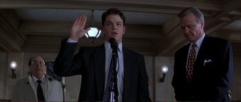 Movie still from “The Rainmaker” (1997), directed by Francis Ford Coppola – A man in a suit is holding up his hand while standing at a microphone; Medium shot, Low angle