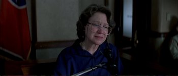 Movie still from “The Rainmaker” (1997), directed by Francis Ford Coppola – An older woman wearing glasses is sitting in front of a microphone; Close Up shot, Over the shoulder angle