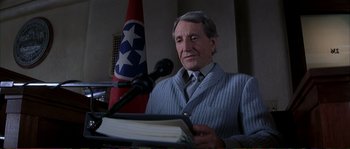 Movie still from “The Rainmaker” (1997), directed by Francis Ford Coppola – An older man is holding a book while standing at a podium; Medium shot, Low angle