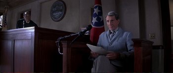 Movie still from “The Rainmaker” (1997), directed by Francis Ford Coppola – A man sitting in front of microphones in front of a flag; Medium shot, Low angle