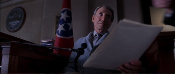 Movie still from “The Rainmaker” (1997), directed by Francis Ford Coppola – A man sitting at a podium in front of microphones; Close Up shot, Low angle