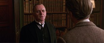 Movie still from “The Remains of the Day” (1993), directed by James Ivory – An older man wearing a suit and tie in front of a book shelf; Medium shot, Over the shoulder angle