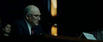 Movie still from “The Report” (2019), directed by Scott Z. Burns – An older man is speaking at a microphone in front of a crowd; Close Up shot, Over the shoulder angle