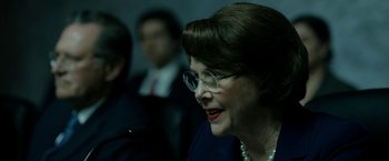 Movie still from “The Report” (2019), directed by Scott Z. Burns – An older woman wearing glasses and a pearl necklace; Close Up shot, Over the shoulder angle
