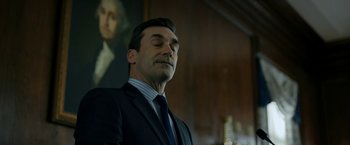 Movie still from “The Report” (2019), directed by Scott Z. Burns – A man wearing a suit and tie in front of a painting; Medium shot, Low angle