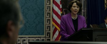Movie still from “The Report” (2019), directed by Scott Z. Burns – A woman in a purple suit stands in front of an american flag; Medium shot, Over the shoulder angle