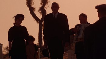 Movie still from “The Right Stuff” (1983), directed by Philip Kaufman – An older man in a suit and tie with a young boy and a woman in a white dress and a man in a black suit; Wide shot, Low angle