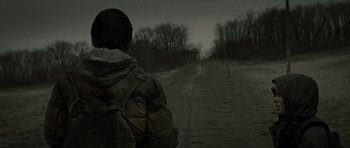 Movie still from “The Road” (2009), directed by John Hillcoat – A person with a backpack walking down a dirt road; Medium shot, Over the shoulder angle