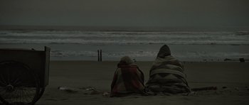 Movie still from “The Road” (2009), directed by John Hillcoat – Two people sitting on the beach under a blanket; Extreme Wide shot, High angle