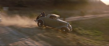 Movie still from “The Rocketeer” (1991), directed by Joe Johnston – An old car driving down a dirt road with a man sitting in the back seat; Wide shot, High angle