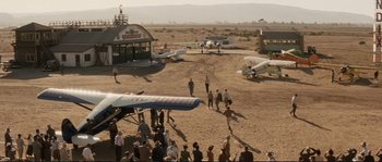 Movie still from “The Rocketeer” (1991), directed by Joe Johnston – A group of people standing in the dirt near airplanes; Extreme Wide shot, High angle