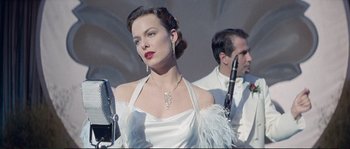 Movie still from “The Rocketeer” (1991), directed by Joe Johnston – A woman in a white dress with a microphone and other people in the background; Medium shot, Low angle