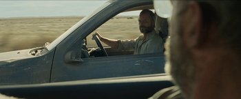 Movie still from “The Rover” (2014), directed by David Michôd – A man sitting in the driver's seat of a car driving down a road; Medium shot, Over the shoulder angle