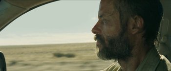 Movie still from “The Rover” (2014), directed by David Michôd – A man with a long beard looking out a window; Close Up shot, Over the shoulder angle