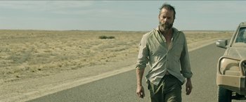Movie still from “The Rover” (2014), directed by David Michôd – A man walking down the side of a road; Medium shot, Over the shoulder angle