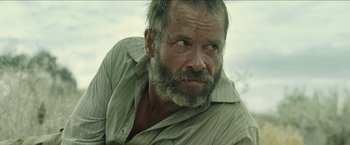 Movie still from “The Rover” (2014), directed by David Michôd – A man with a beard and a shirt is looking at the camera; Close Up shot, Low angle