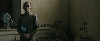Movie still from “The Rover” (2014), directed by David Michôd – A man standing next to another man sitting in a room; Medium shot, Low angle