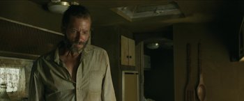 Movie still from “The Rover” (2014), directed by David Michôd – A man standing in a room next to an open door; Medium shot, Low angle