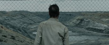 Movie still from “The Rover” (2014), directed by David Michôd – A man standing in front of a fence looking out at the ocean; Wide shot, High angle