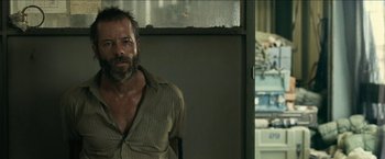 Movie still from “The Rover” (2014), directed by David Michôd – A man standing in front of a wall with cars in the background; Medium shot, Over the shoulder angle