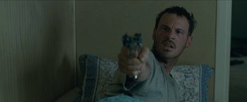 Movie still from “The Rover” (2014), directed by David Michôd – A man is holding a gun in his hand while sitting on a couch; Close Up shot, Low angle