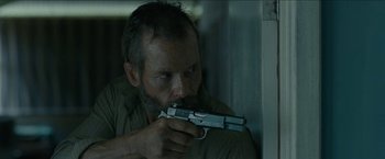 Movie still from “The Rover” (2014), directed by David Michôd – A man holding a gun while sitting in a room; Close Up shot, Low angle