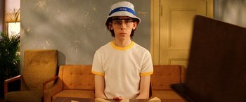 Movie still from “The Royal Tenenbaums” (2001), directed by Wes Anderson – A young man wearing glasses and a hat sitting at a table; Medium shot, Low angle
