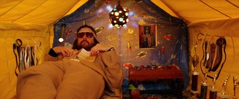 Movie still from “The Royal Tenenbaums” (2001), directed by Wes Anderson – A man with a beard and sunglasses is sitting in a tent; Medium shot, Overhead angle